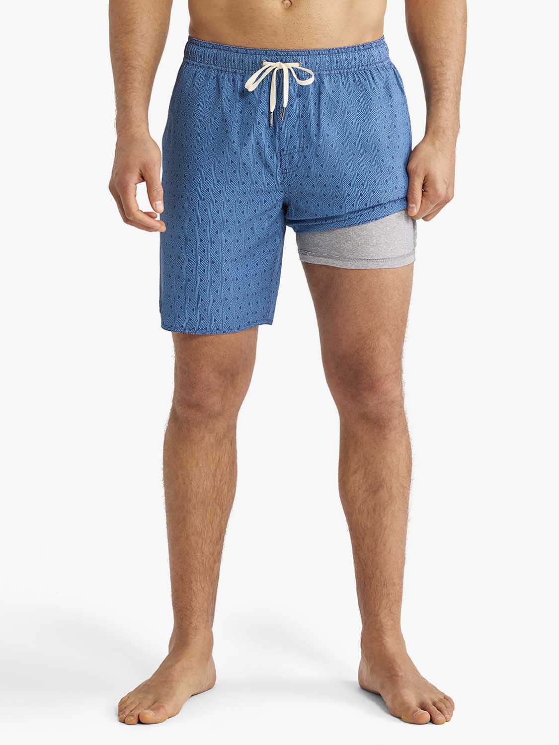 Man wearing electric blue geometric patterned swim trunk with white drawstring and gray underlay.