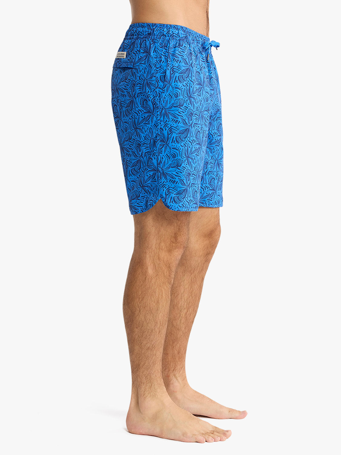 Man wearing blue swim shorts with dark leaf pattern print and drawstring waistband.