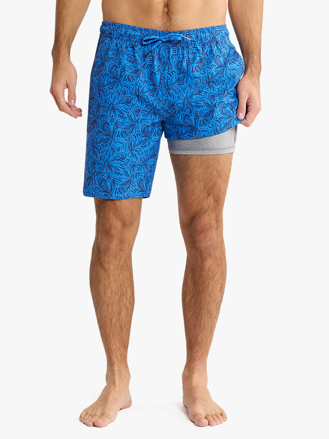 Man wearing blue swim trunks with dark leaf print pattern and gray waistband detail.
