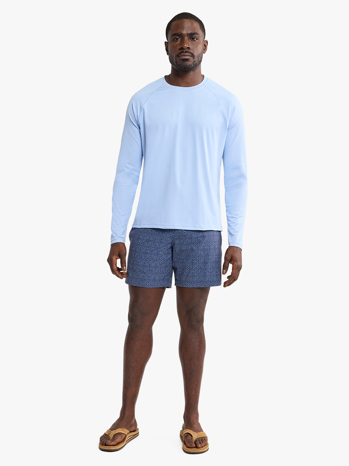 Man wearing a light blue long-sleeve rashguard with dark patterned swim shorts and tan sandals.