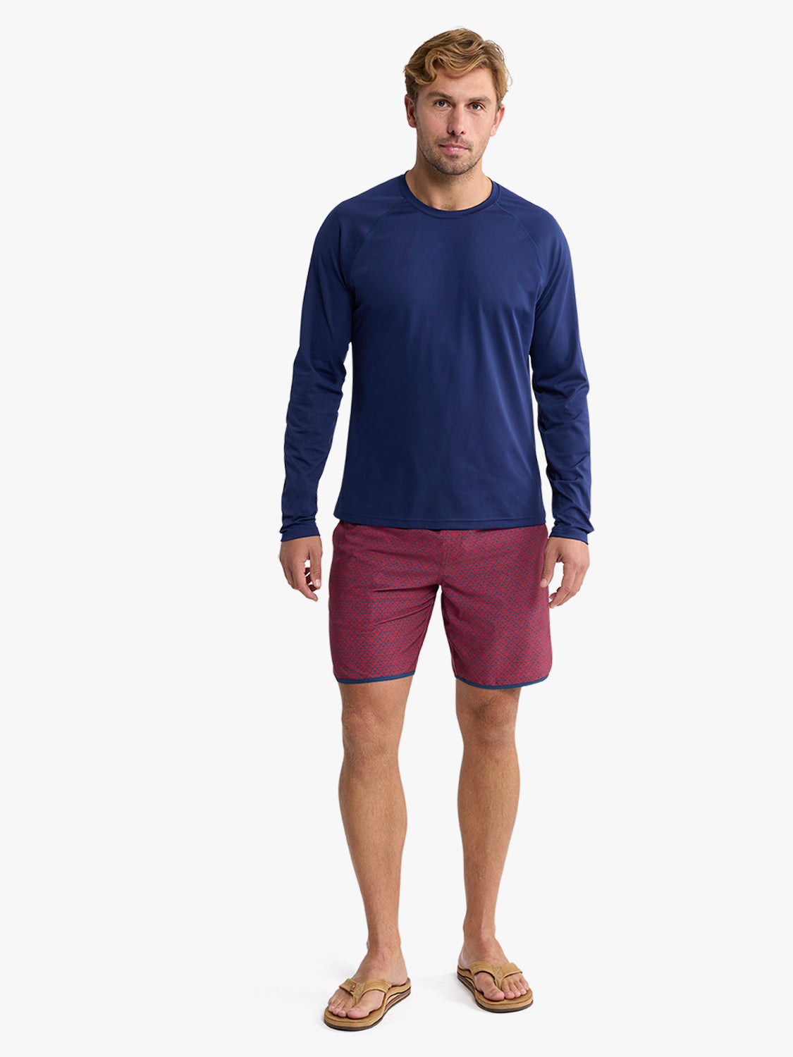 Man wearing a navy long-sleeve rashguard with burgundy patterned swim shorts and sandals.