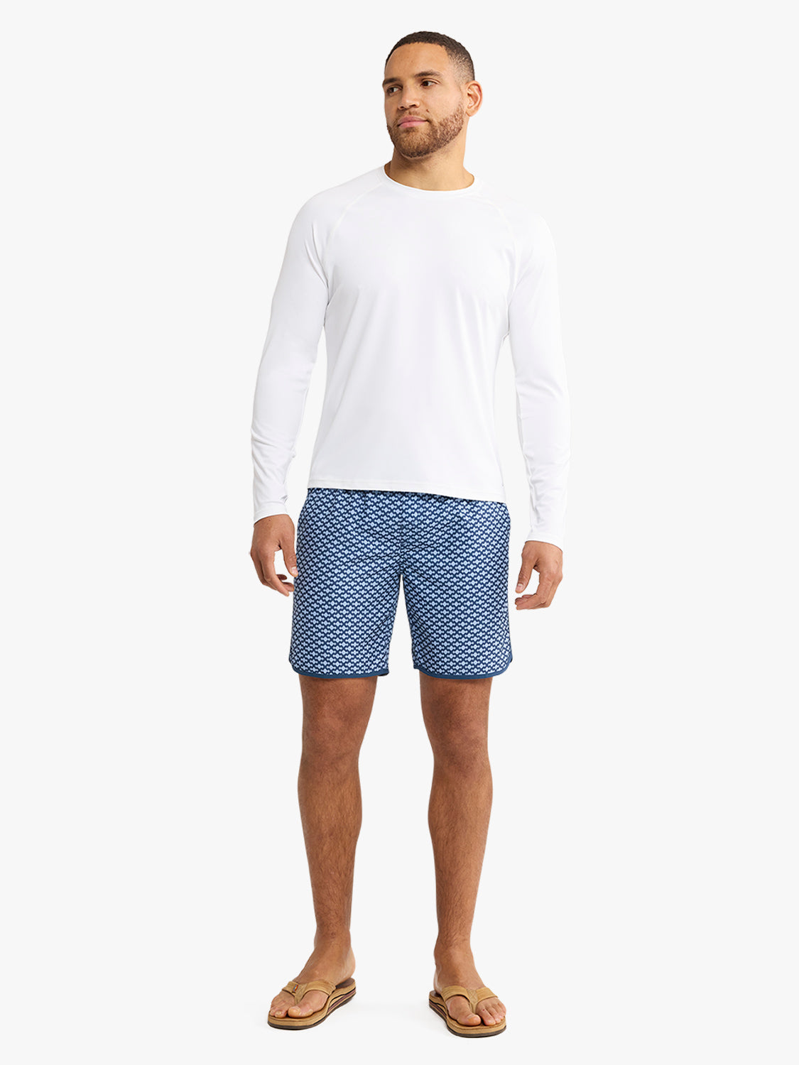 Man wearing white long-sleeve rashguard with blue patterned swim shorts and tan sandals.
