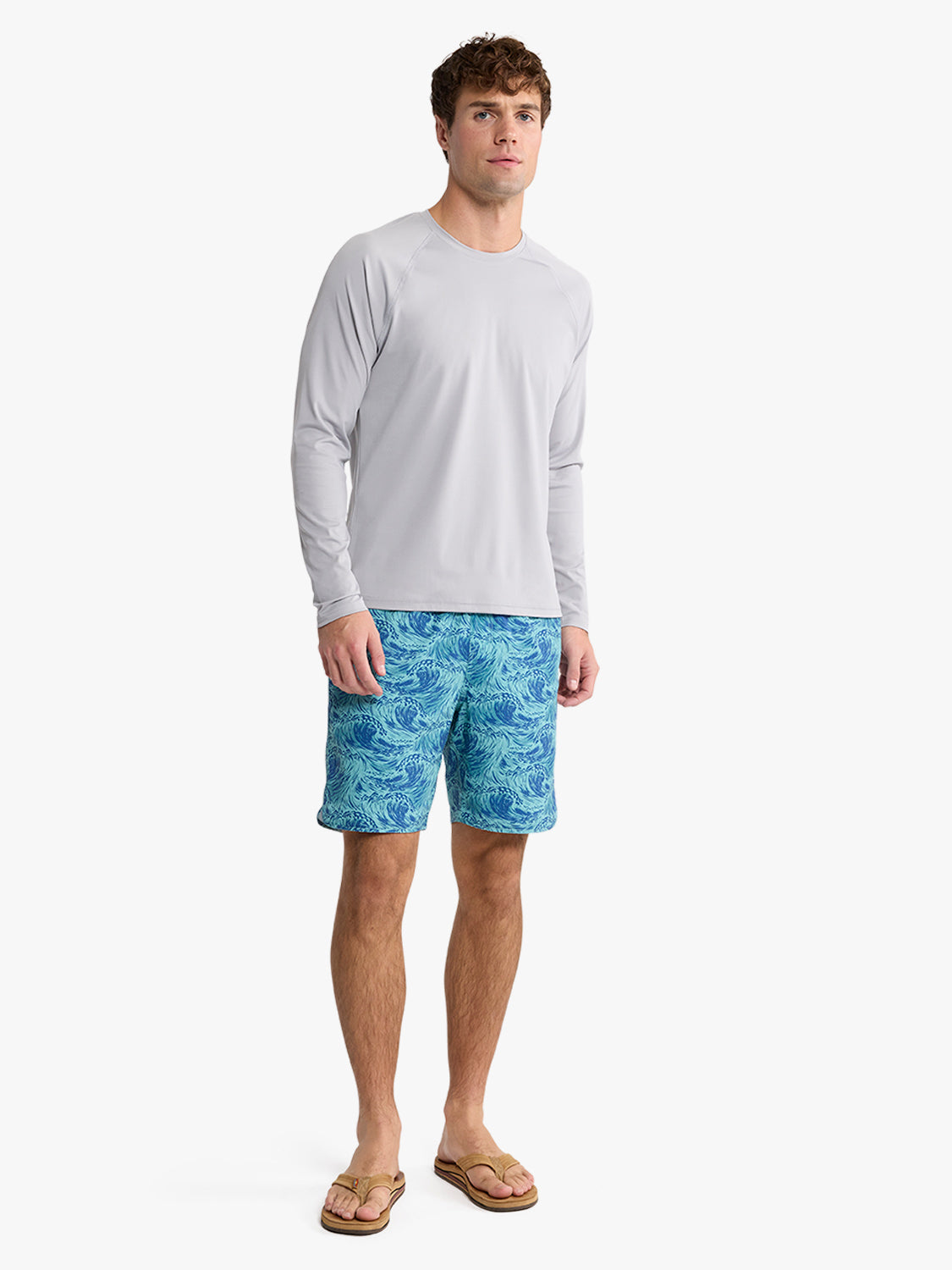 Man wearing a gray long-sleeve rashguard with blue patterned swim shorts and tan sandals.