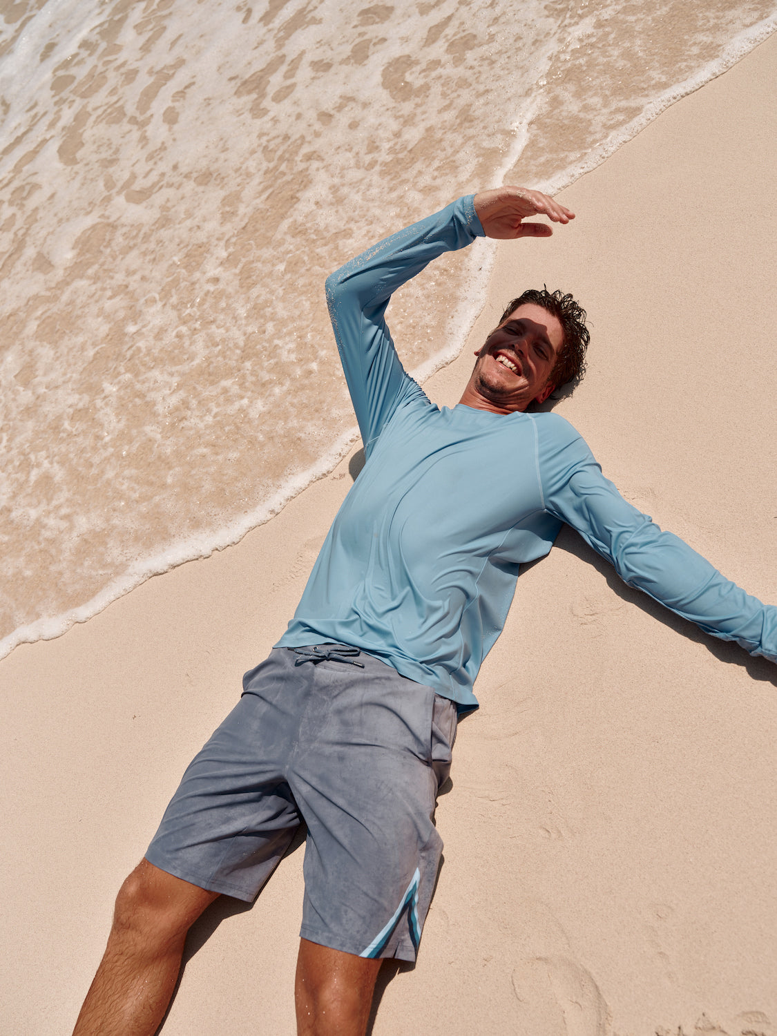 Man laying in the sand on the beach wearing the anchor light blue rashguard and ozone. 