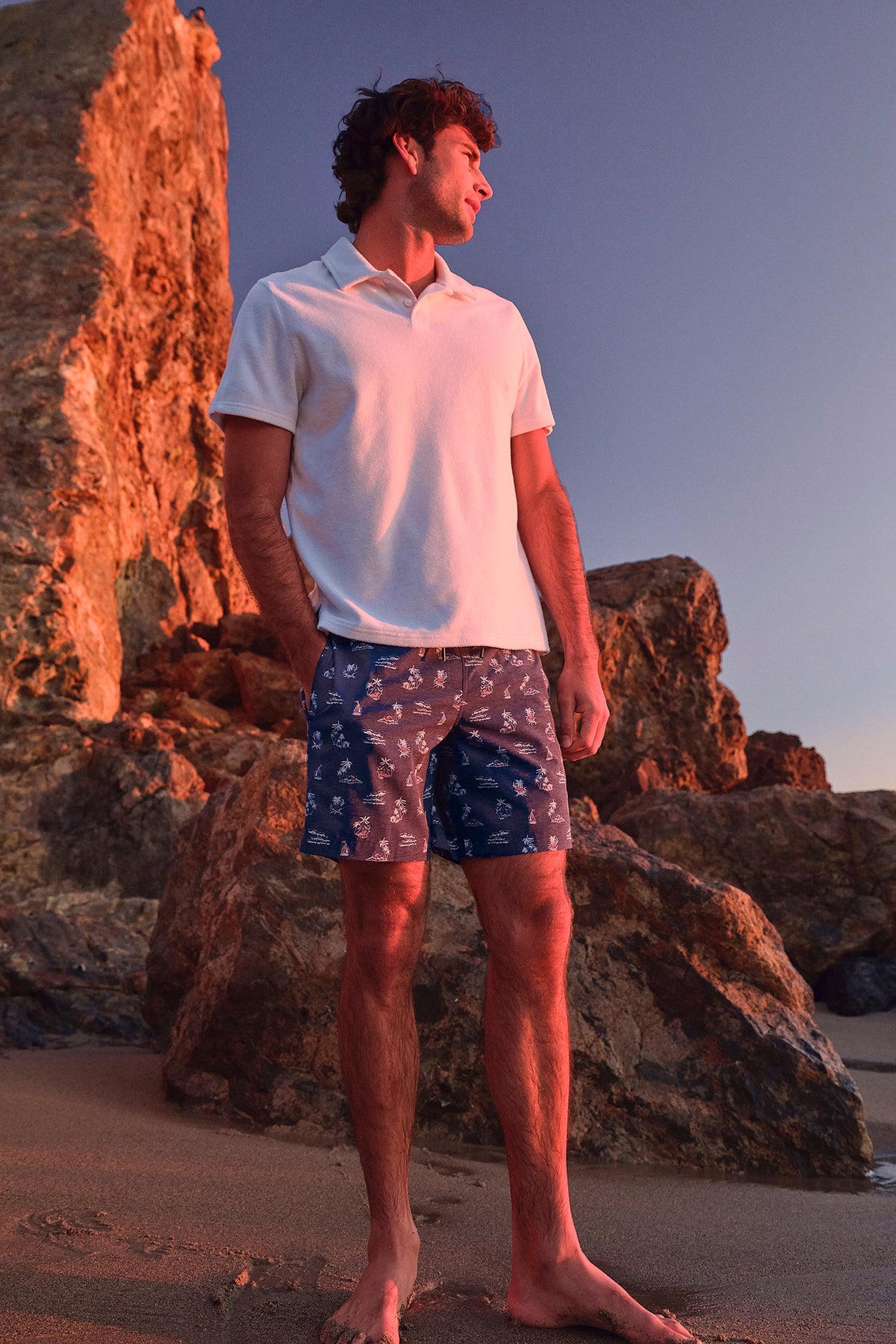 Man wearing light pink short-sleeve polo shirt and navy patterned swim shorts standing on beach.