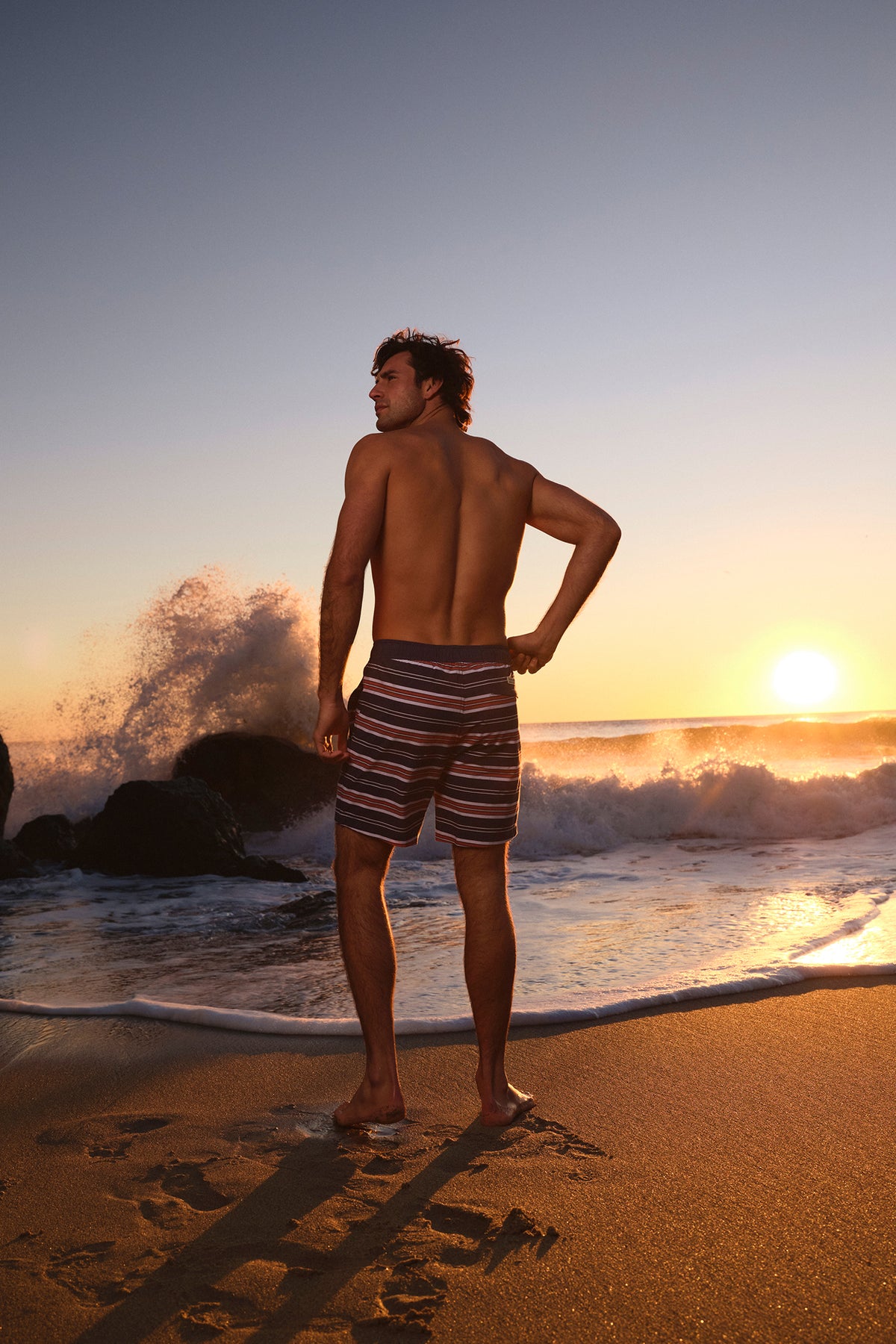 Man wearing striped swim trunks stands on beach at sunset with waves crashing behind him.