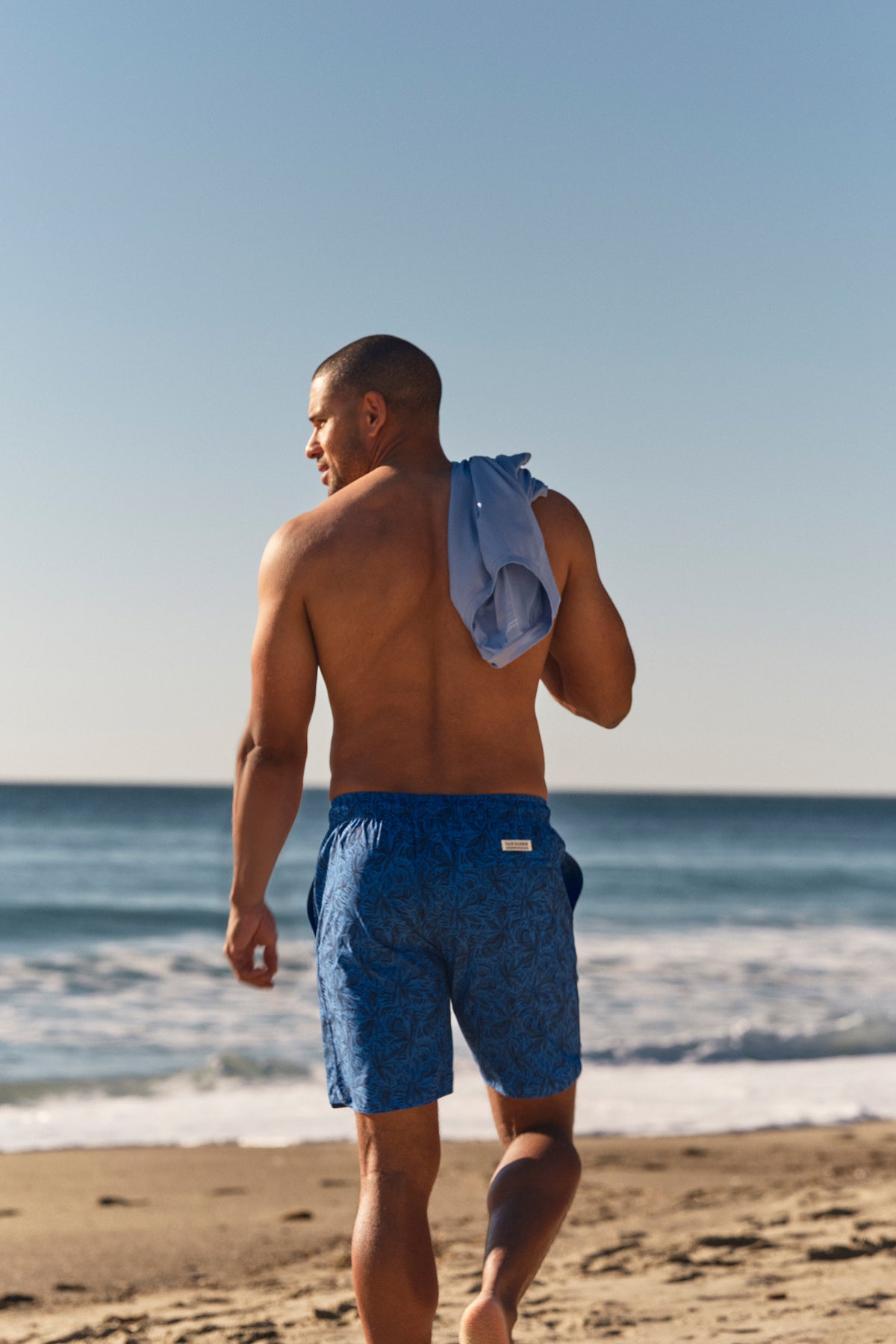 Man wearing blue patterned swim shorts walks on beach carrying towel.