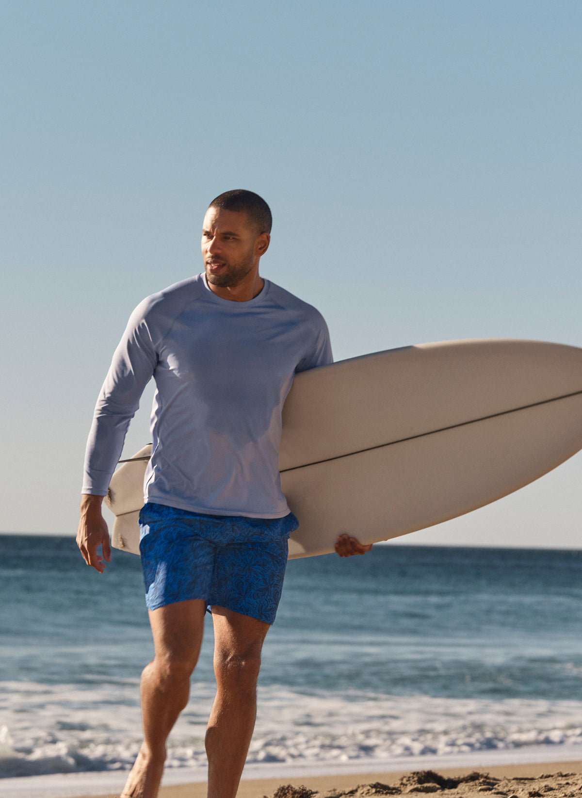 Man wearing turquoise swim trunks with sun and star pattern walking on beach with surfboard.