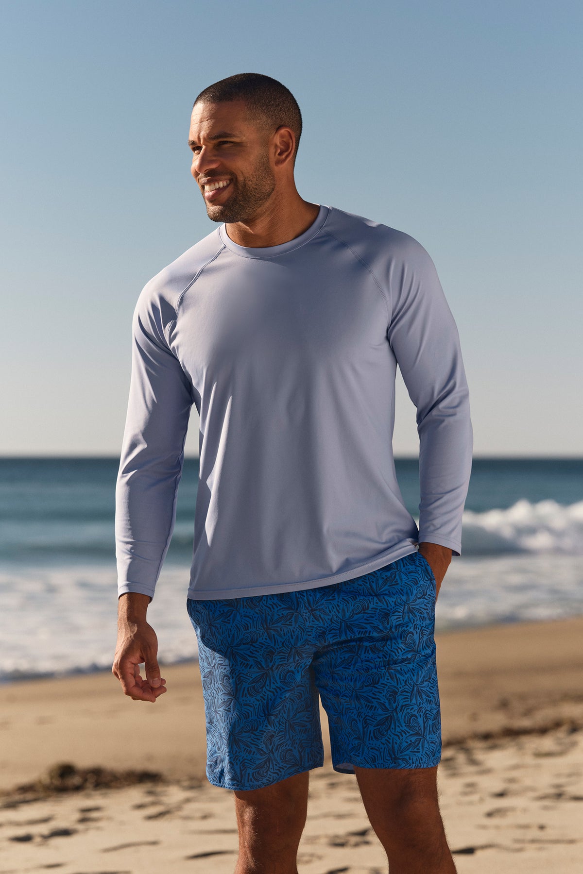 Man wearing lavender long-sleeve shirt and turquoise patterned swim trunks standing on beach.