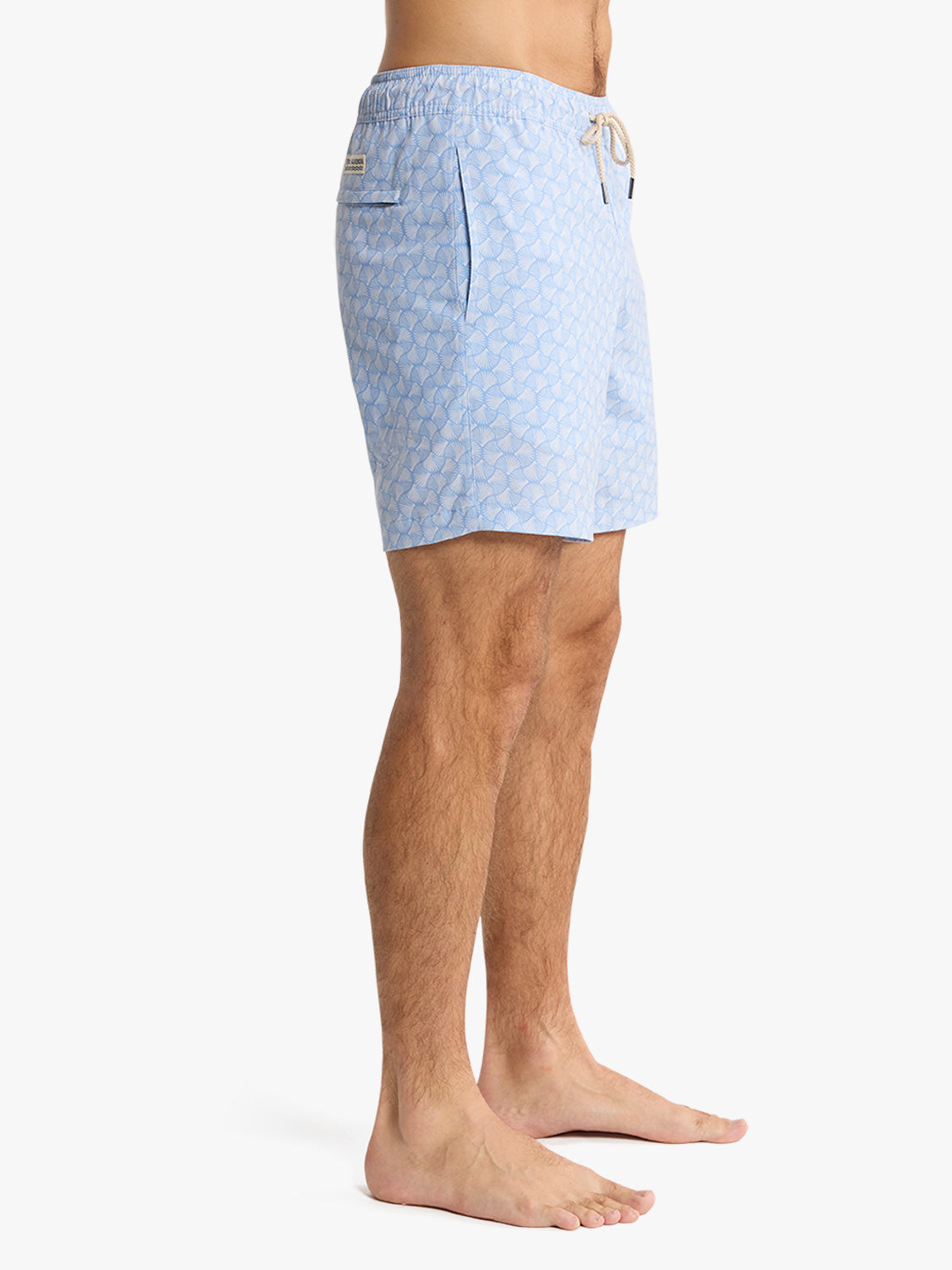 Man wearing light blue swim trunks with a repeating shell pattern and drawstring waistband.