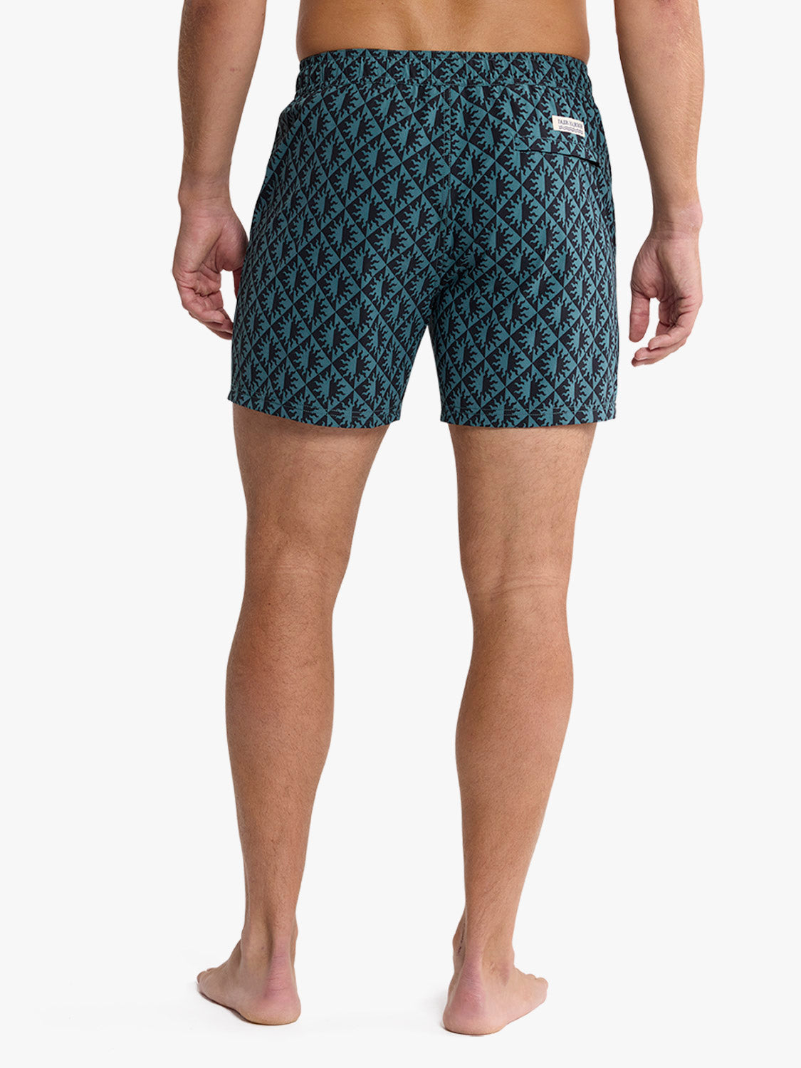 Man wearing teal and navy geometric patterned swim trunks with elastic waistband and small logo patch.