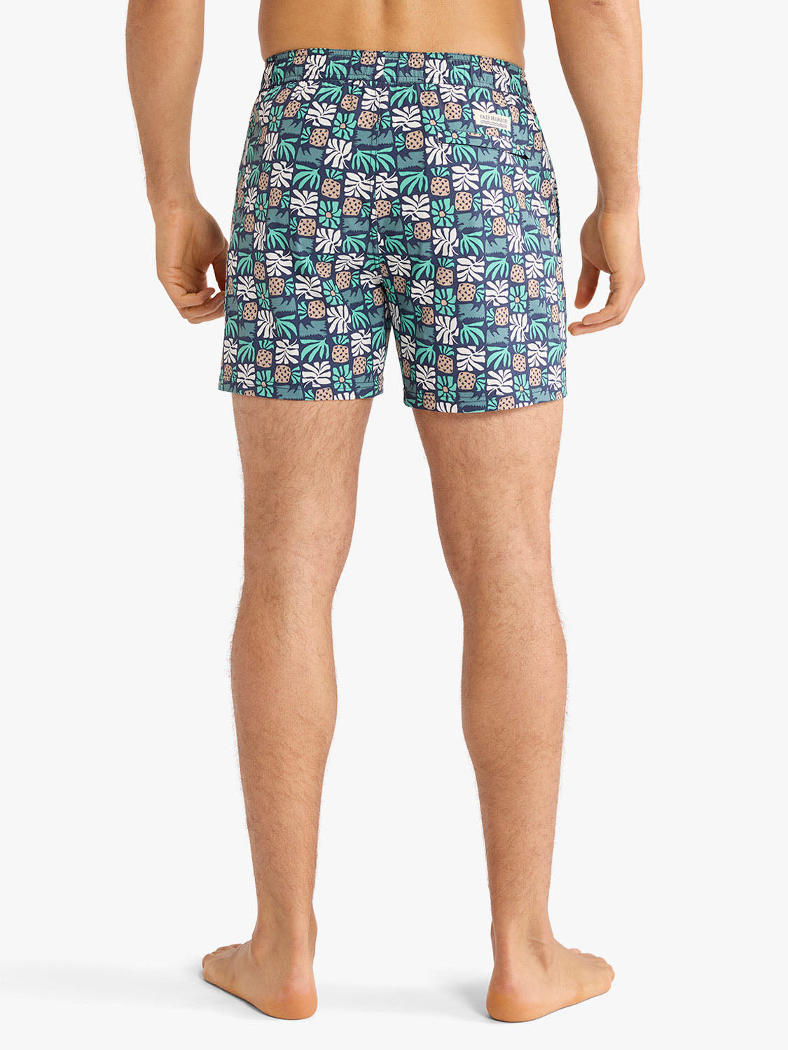 Man wearing navy swim trunks with white and green palm tile pattern shown from behind.