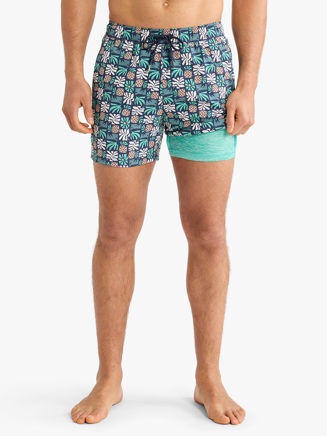 Man wearing navy swim trunks with white and green palm tile pattern and turquoise mesh liner shorts.