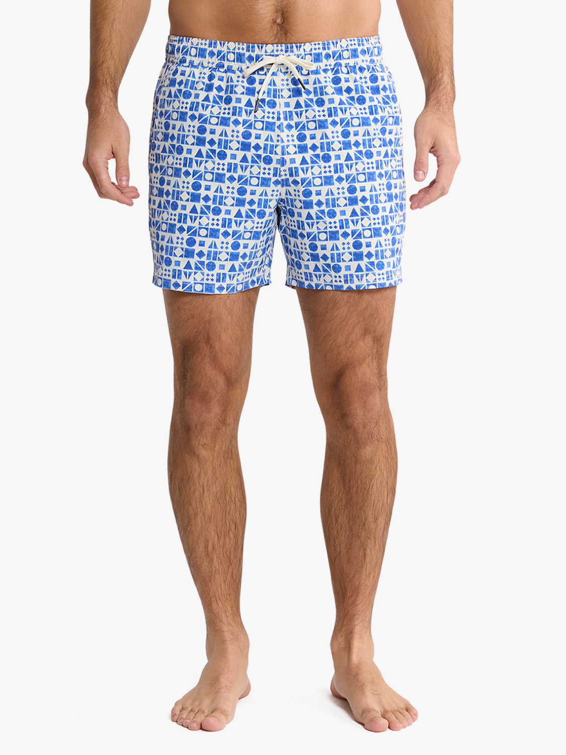 Man wearing blue and white geometric patterned swim trunks standing against a plain background.