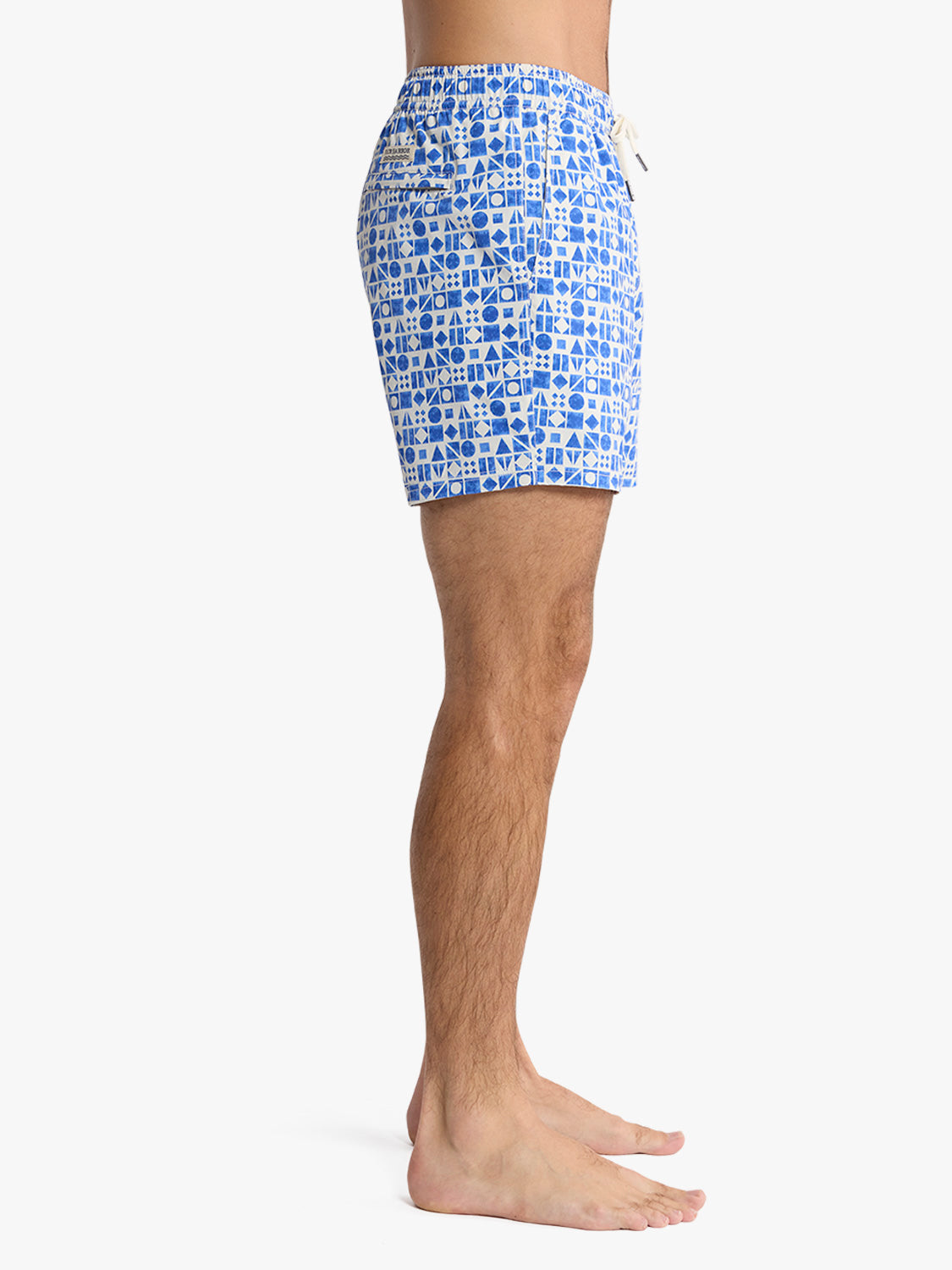 Man wearing blue and white geometric patterned swim trunks standing against neutral background.