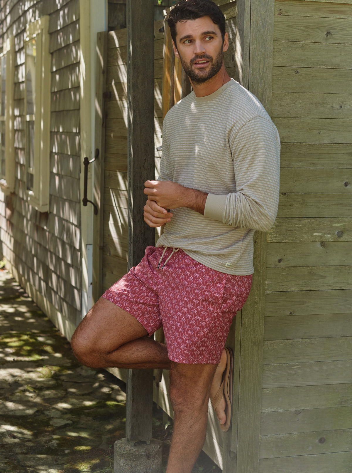 Man wearing a striped long-sleeve shirt and red patterned swim trunk standing outdoors.