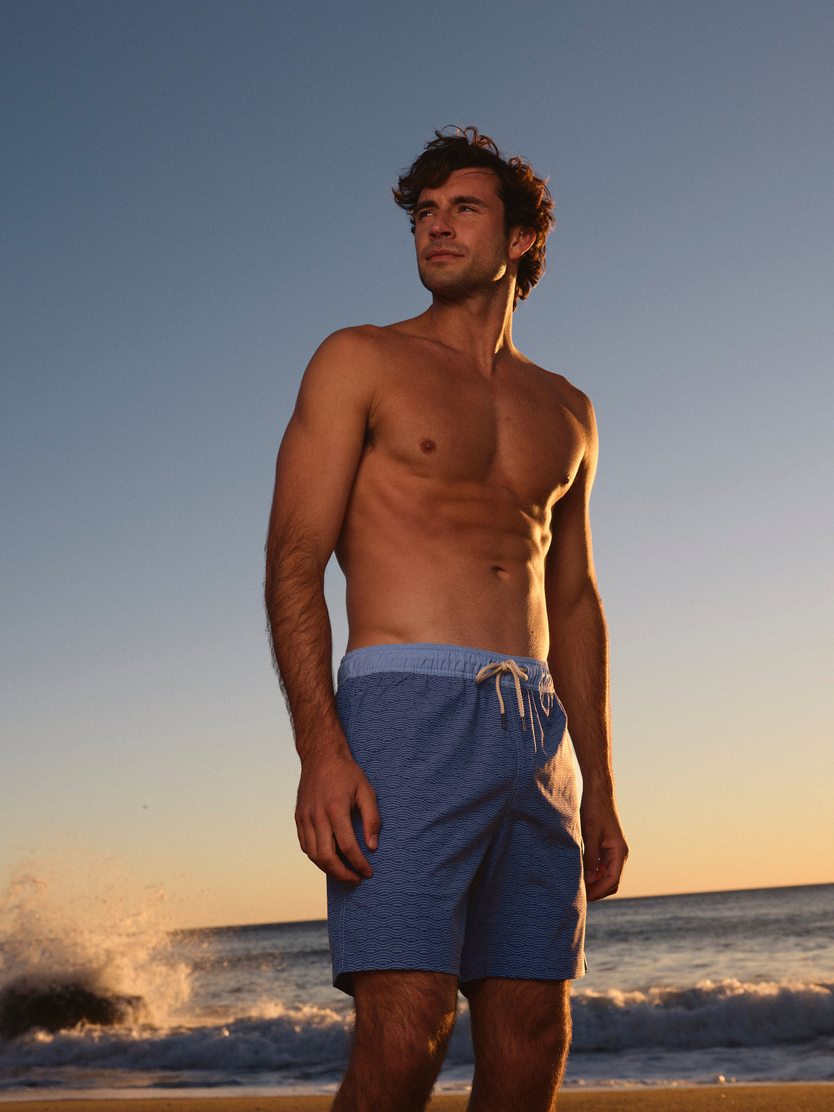 Man wearing blue swim trunks with wave pattern standing on beach at sunset.