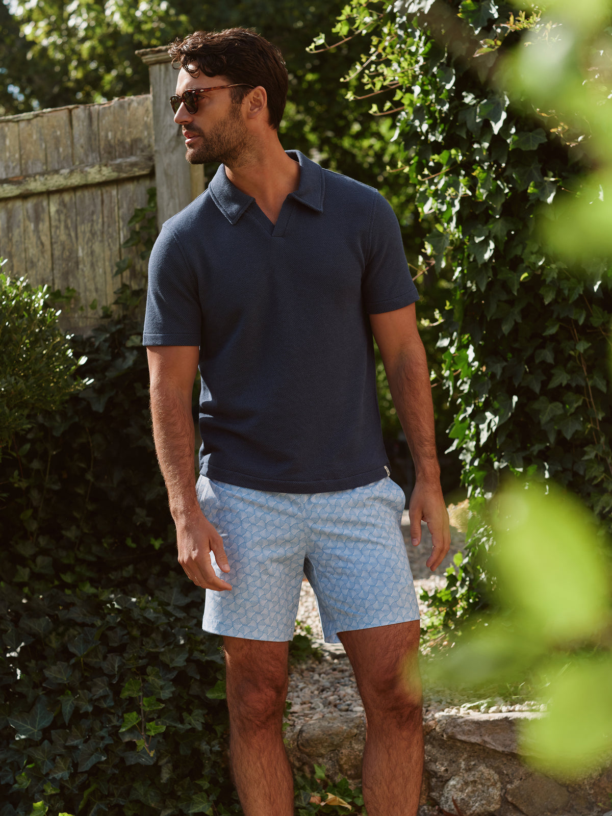 Man wearing navy polo shirt and light blue patterned shorts standing in garden setting.