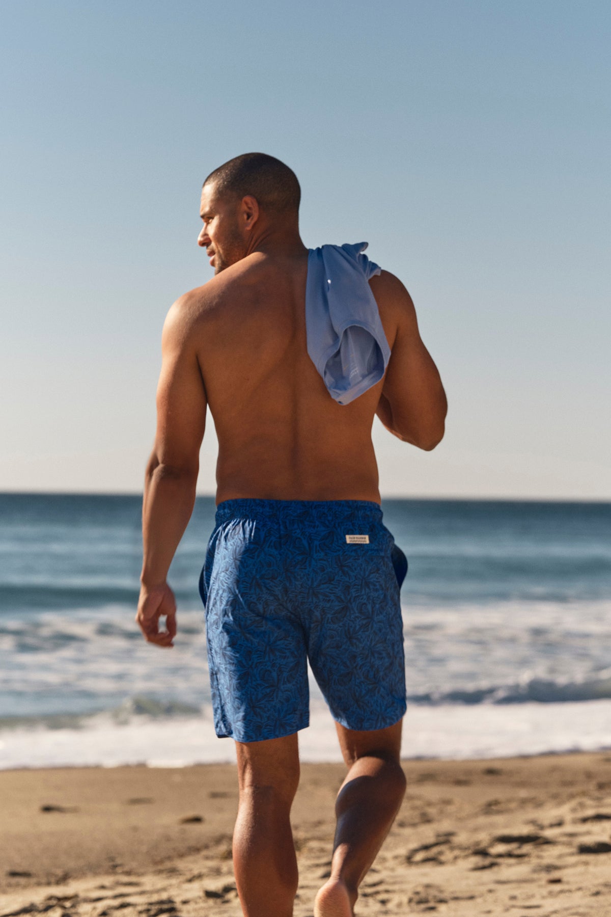 Man wearing island blue leaf-patterned swim shorts and matching towel on a beach.