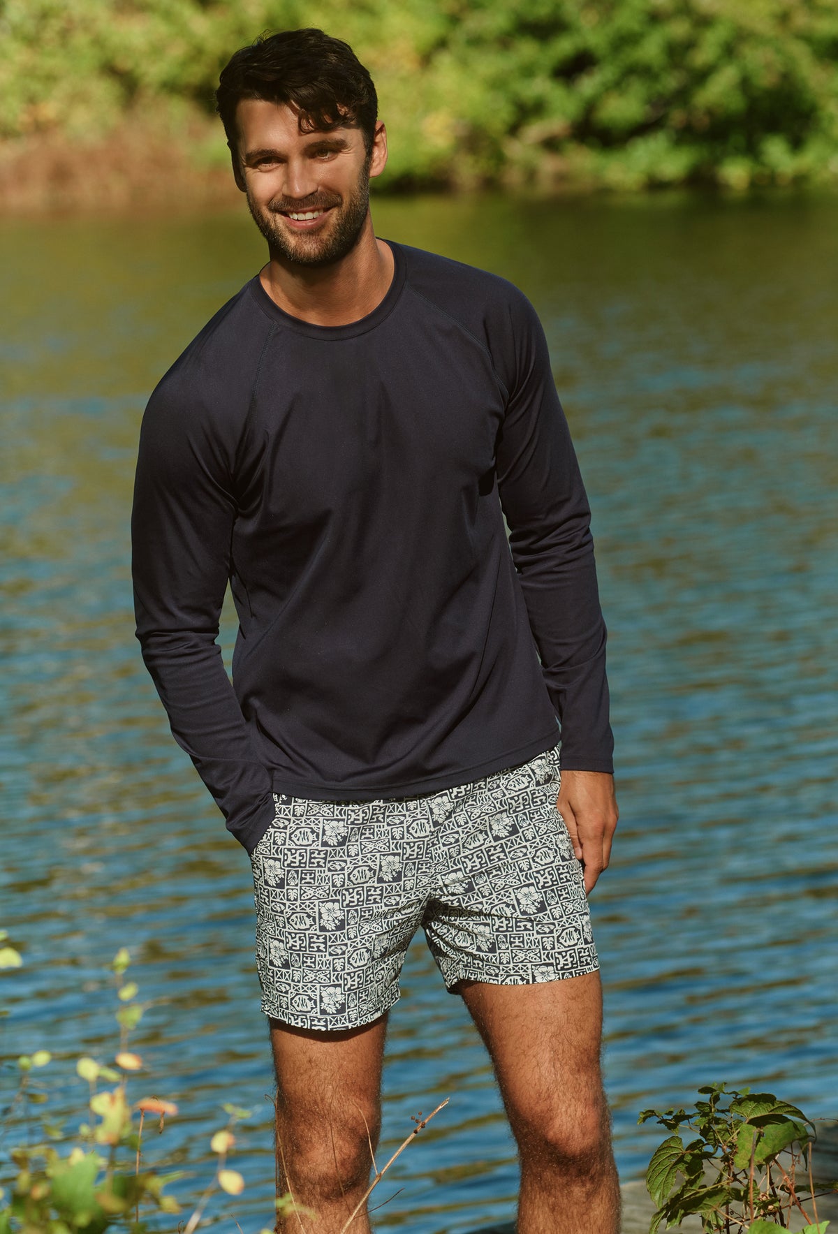 Man wearing black long-sleeve shirt and patterned shorts standing by water.