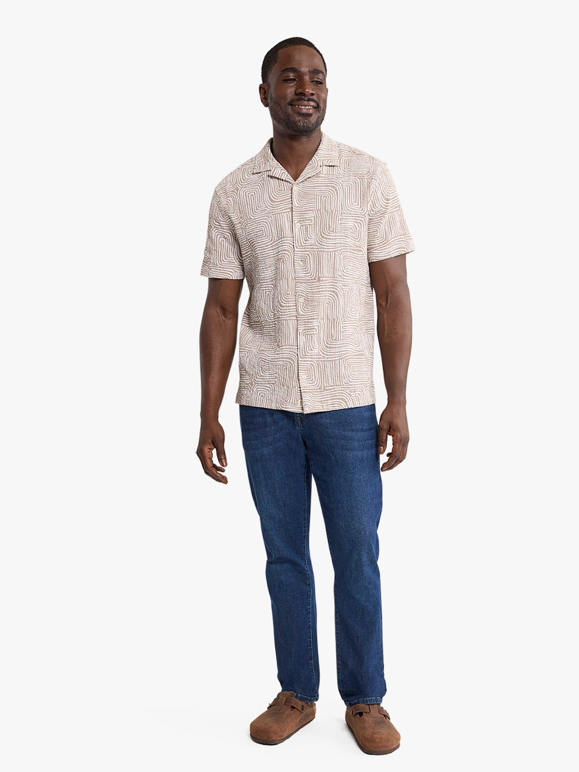 Man wearing a short-sleeved tan camp shirt with abstract wavy pattern and dark blue jeans.