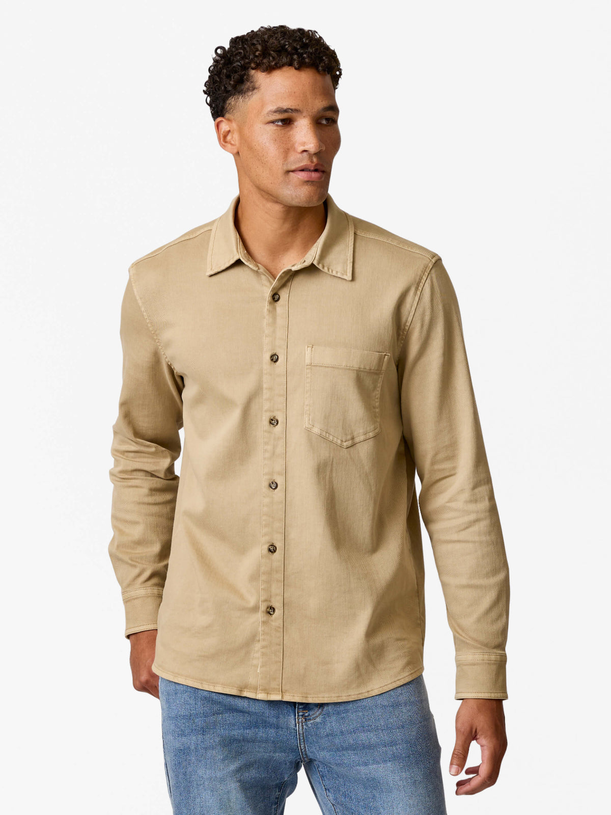 Man wearing a khaki button-up denim shirt with long sleeves and a chest pocket over blue jeans.