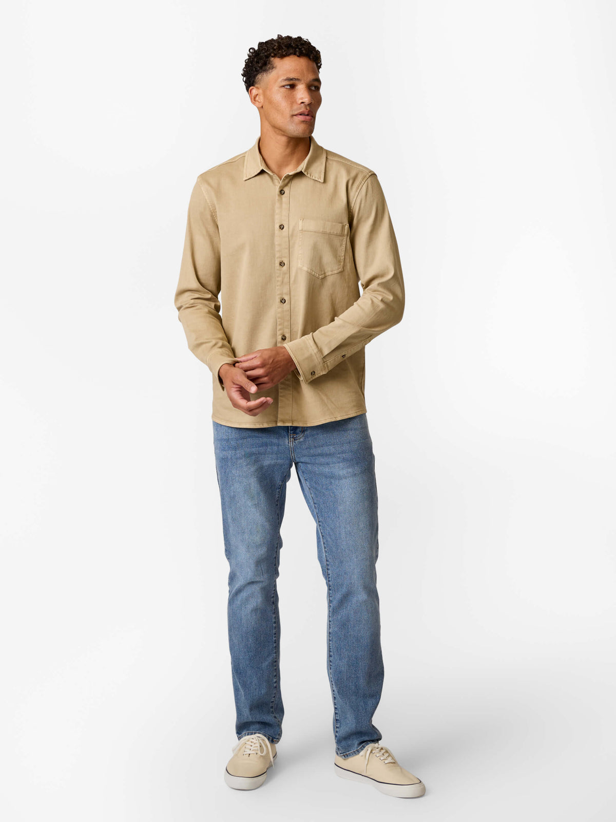Man wearing a khaki denim shirt with long sleeves and chest pocket over blue jeans.