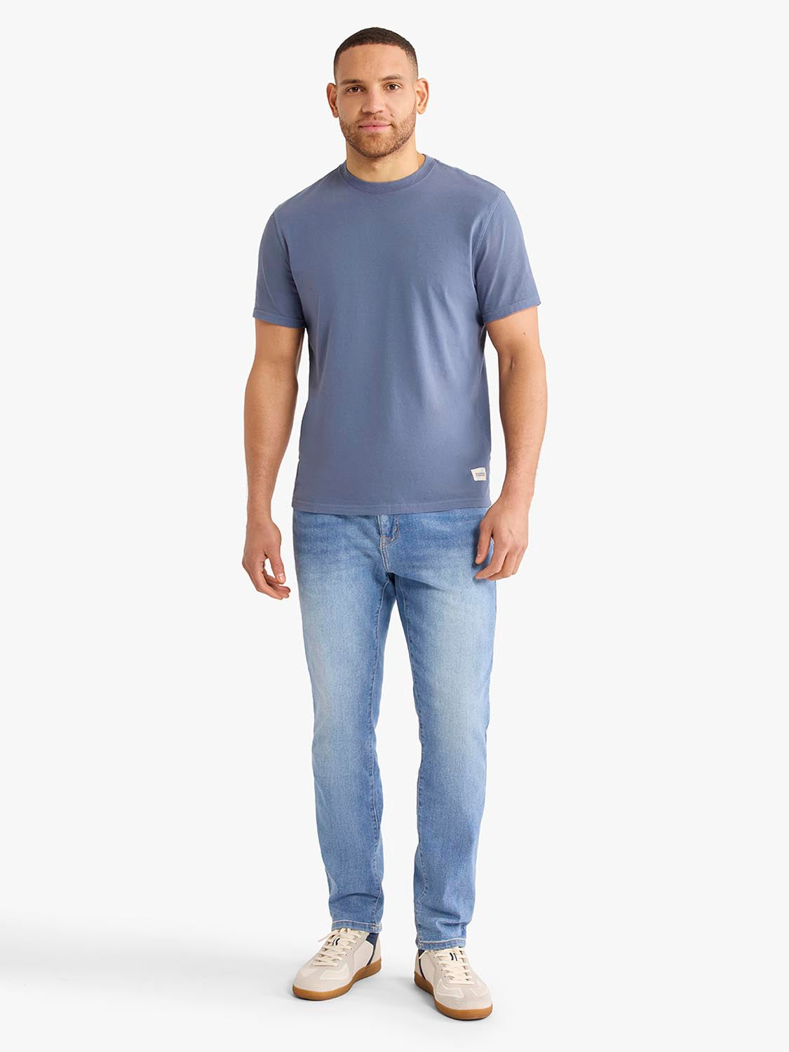 Man wearing a blue-gray t-shirt and light blue beach wash denim pants with white sneakers.