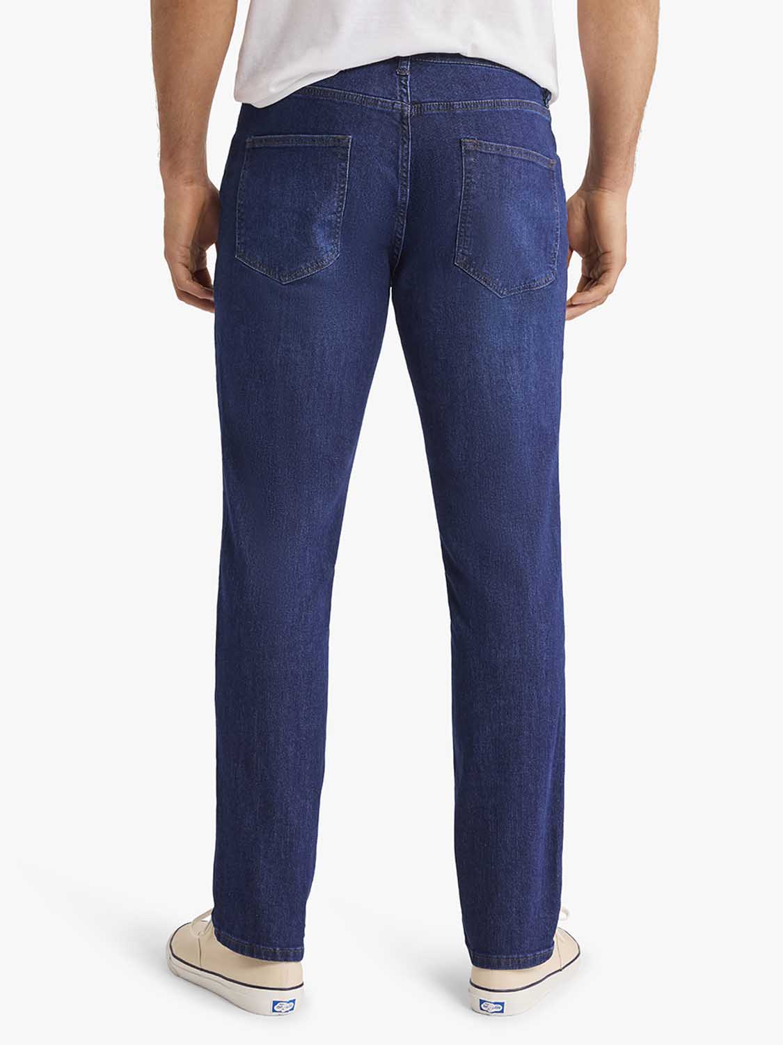 Man wearing dark blue denim pants with back pockets, shown from behind with white shirt and canvas shoes.
