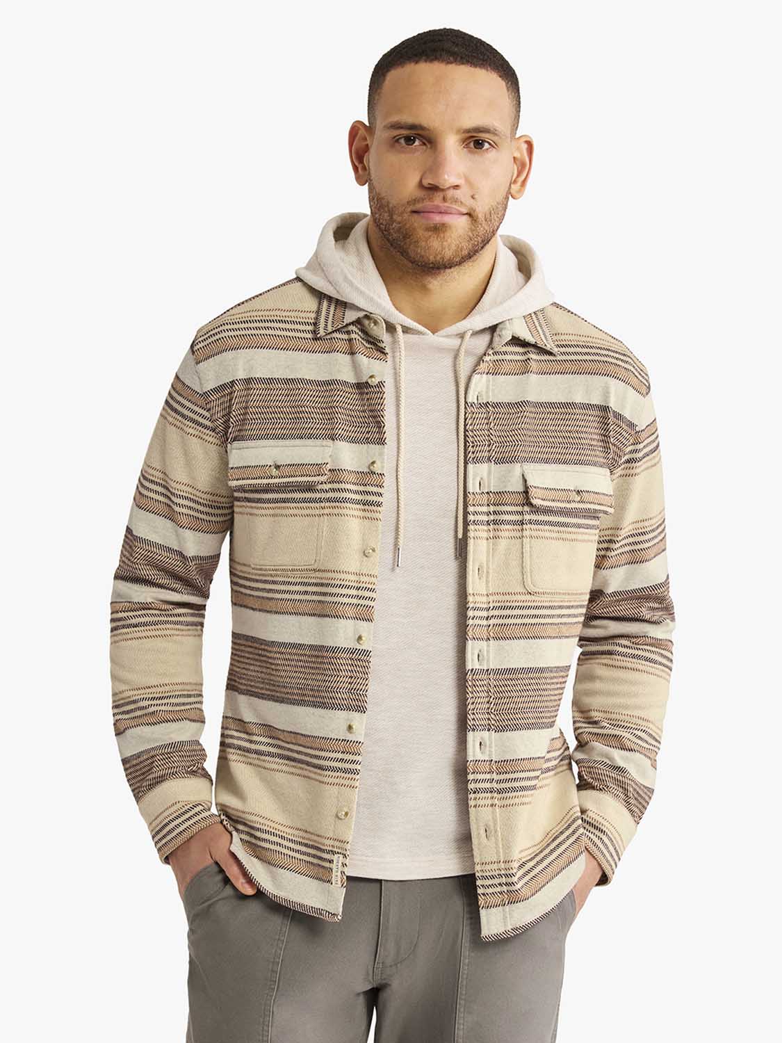 Man wearing a beige and brown striped flannel shirt over a cream hoodie with gray pants.