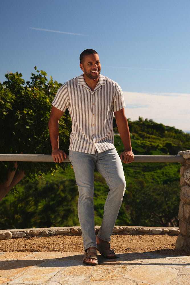 Man wearing light blue stretch denim pants and a striped shirt leaning against a wooden railing outdoors.