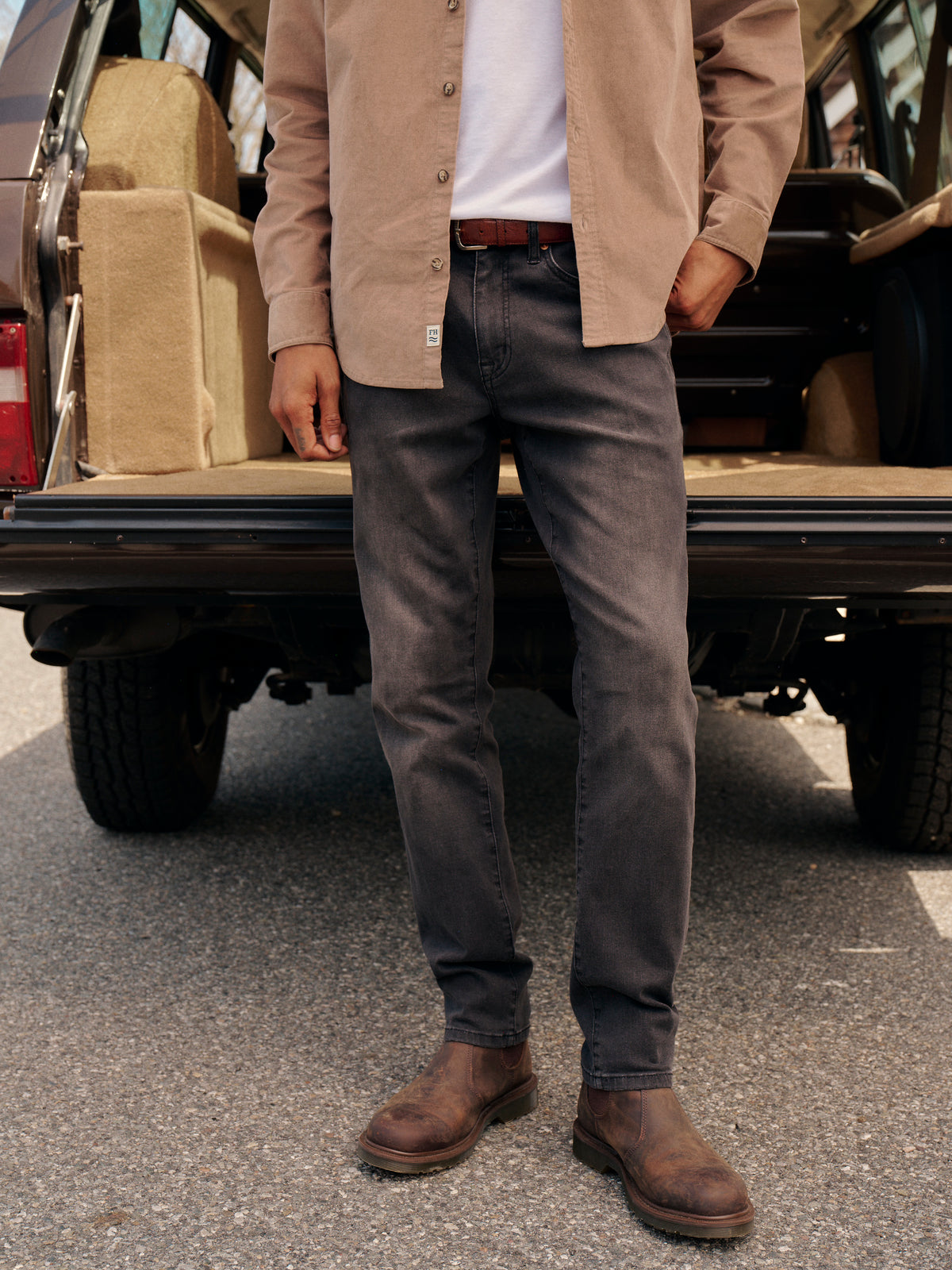 Person wearing black wash denim pants with a tan shirt and brown boots by a truck.