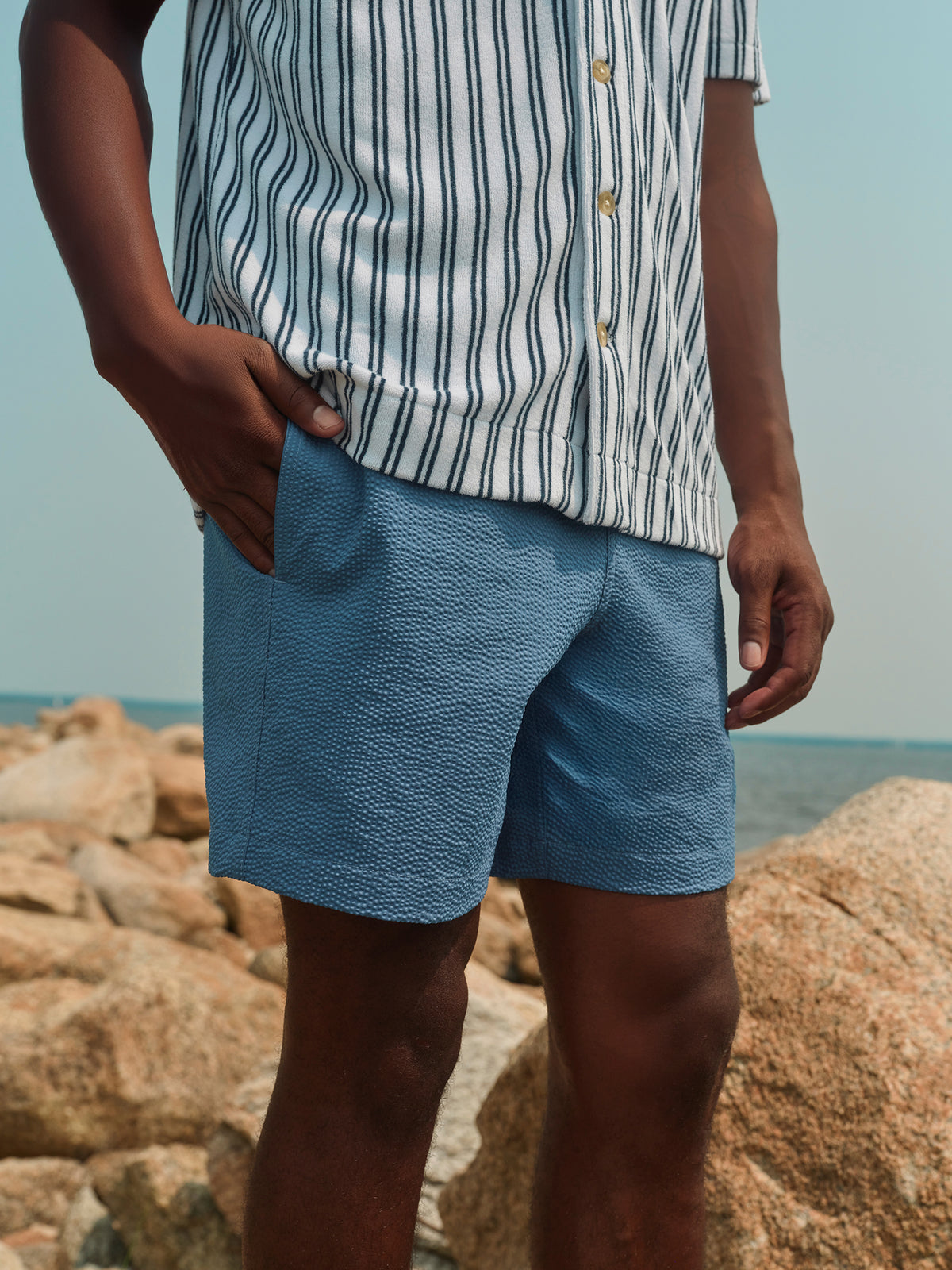 Man wearing striped short sleeve button-up shirt and blue textured shorts standing on beach rocks.