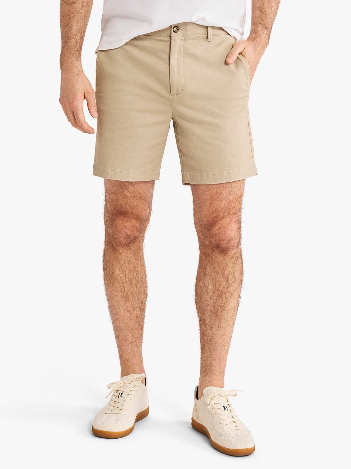 Man wearing khaki shorts with white t-shirt and cream lace-up sneakers standing against white background.