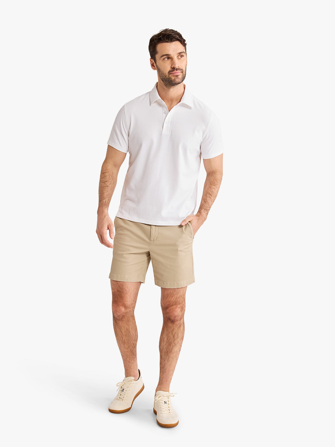 Man wearing white polo shirt and khaki shorts standing against white background.