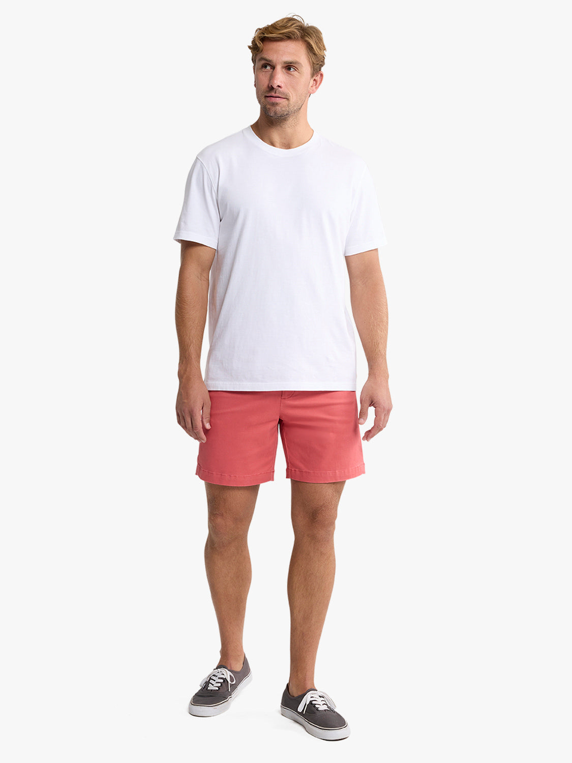 Man wearing white t-shirt and washed red shorts standing against white background.