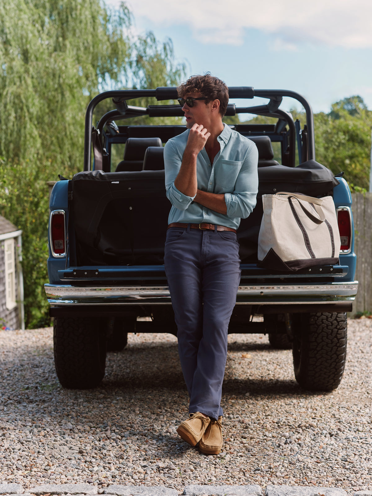 Man wearing navy five-pocket pants and light blue shirt leaning against blue pickup truck bed.