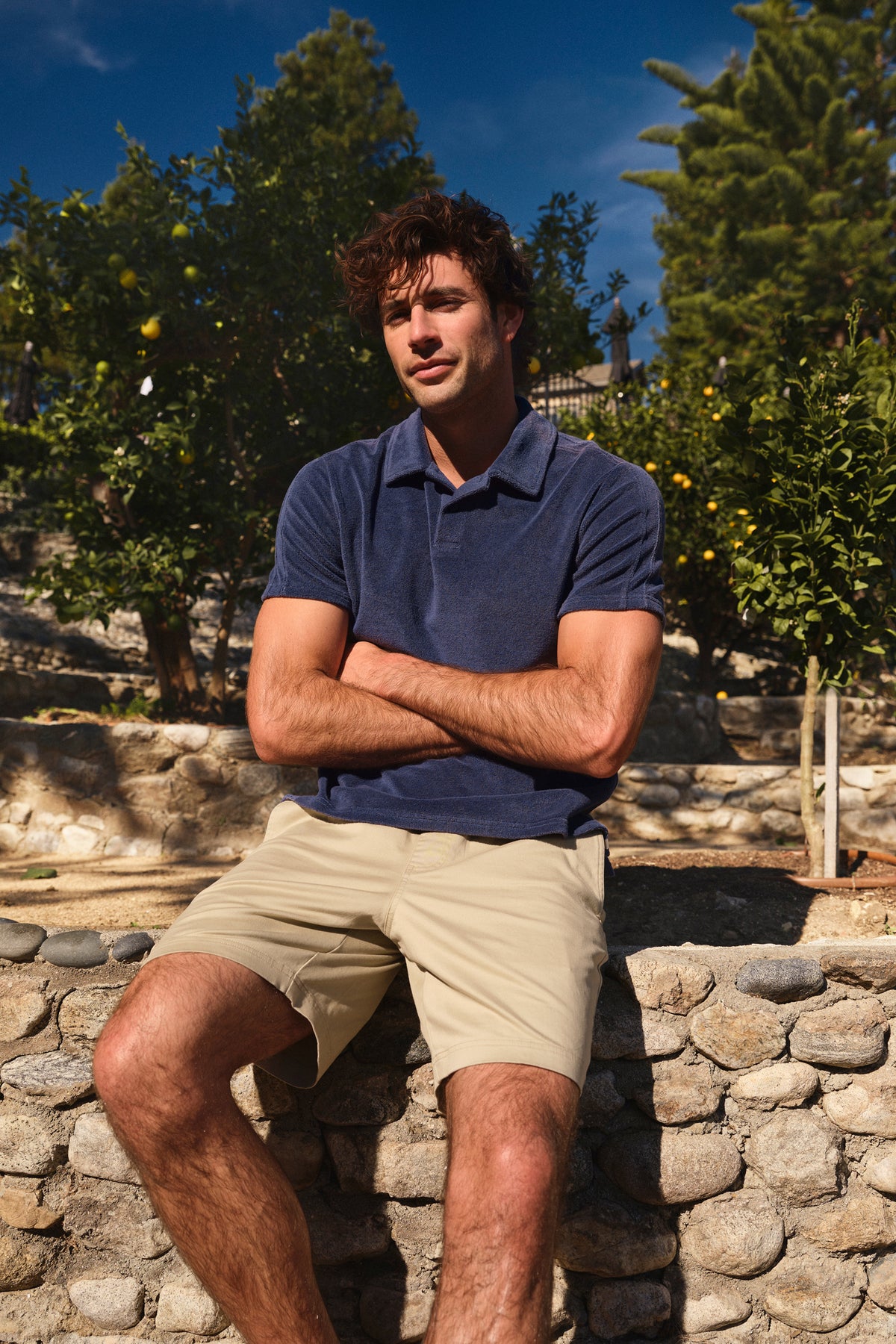 Man wearing a navy polo shirt and khaki pull-on shorts sits on a stone wall in a sunny orchard setting.