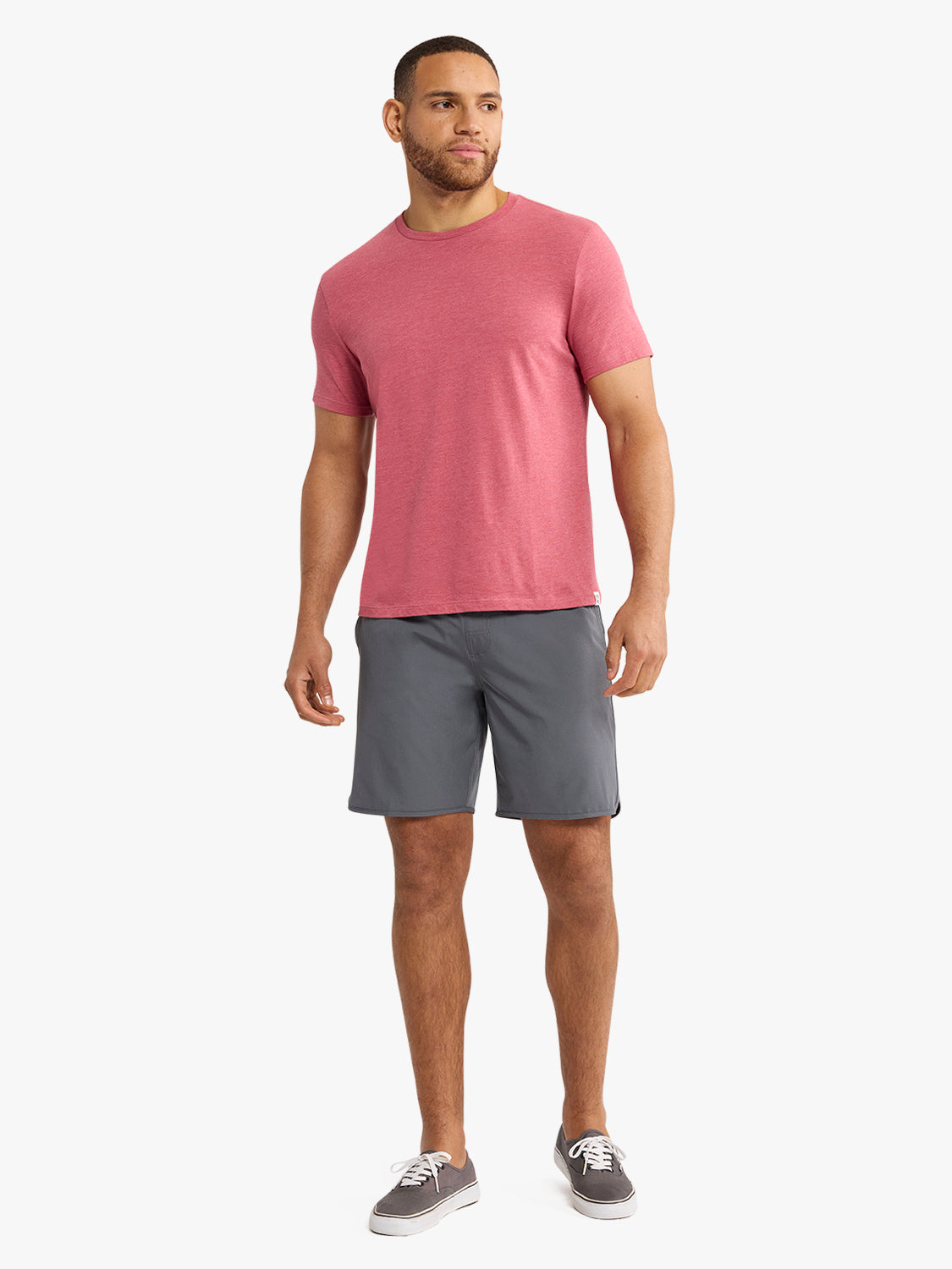 Man wearing a red crew neck t-shirt with gray shorts and sneakers against a white background.