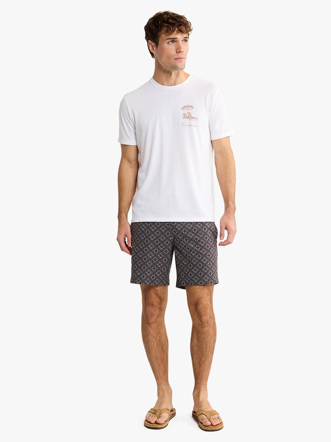 Man wearing a white graphic t-shirt with small chest print and patterned shorts standing against a white background.