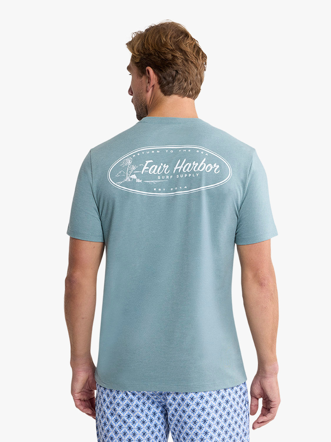 Man wearing a dusty blue short-sleeve graphic tee with white Fair Harbor Surf Supply text and logo on the back.