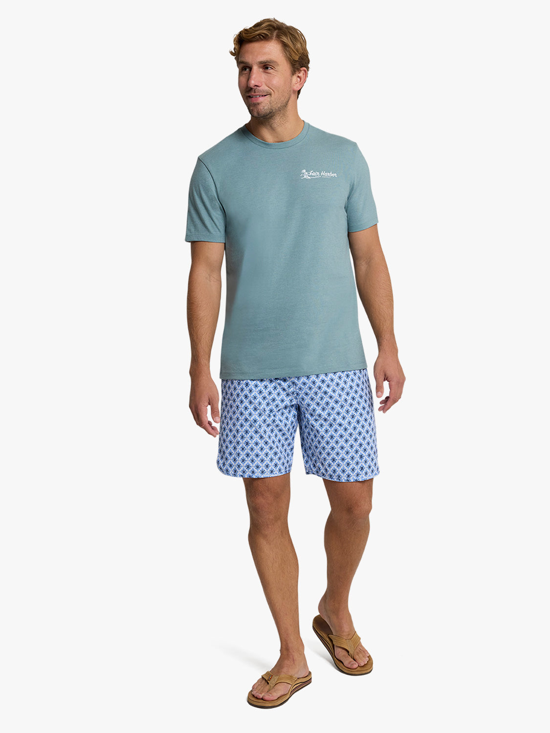 Man wearing a dusty blue graphic tee with white chest logo and patterned blue shorts.