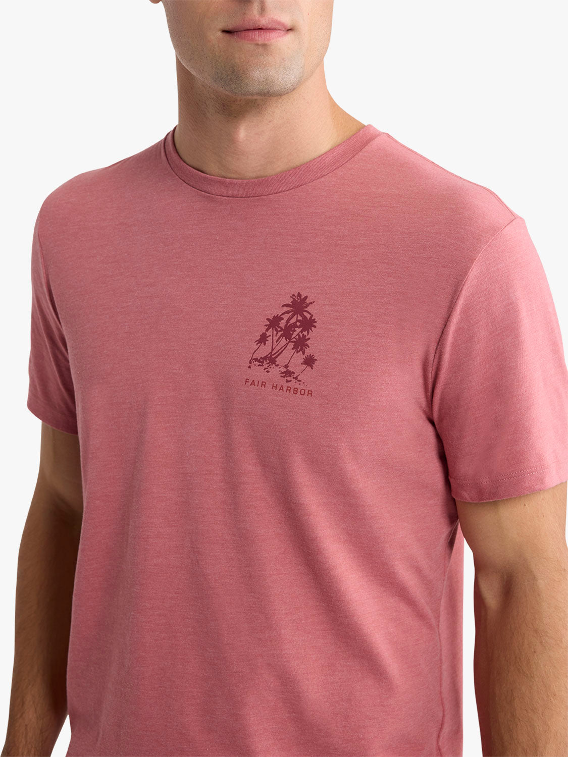 Man wearing a mauve-pink graphic tee with palm tree and Fair Harbor text design on chest.