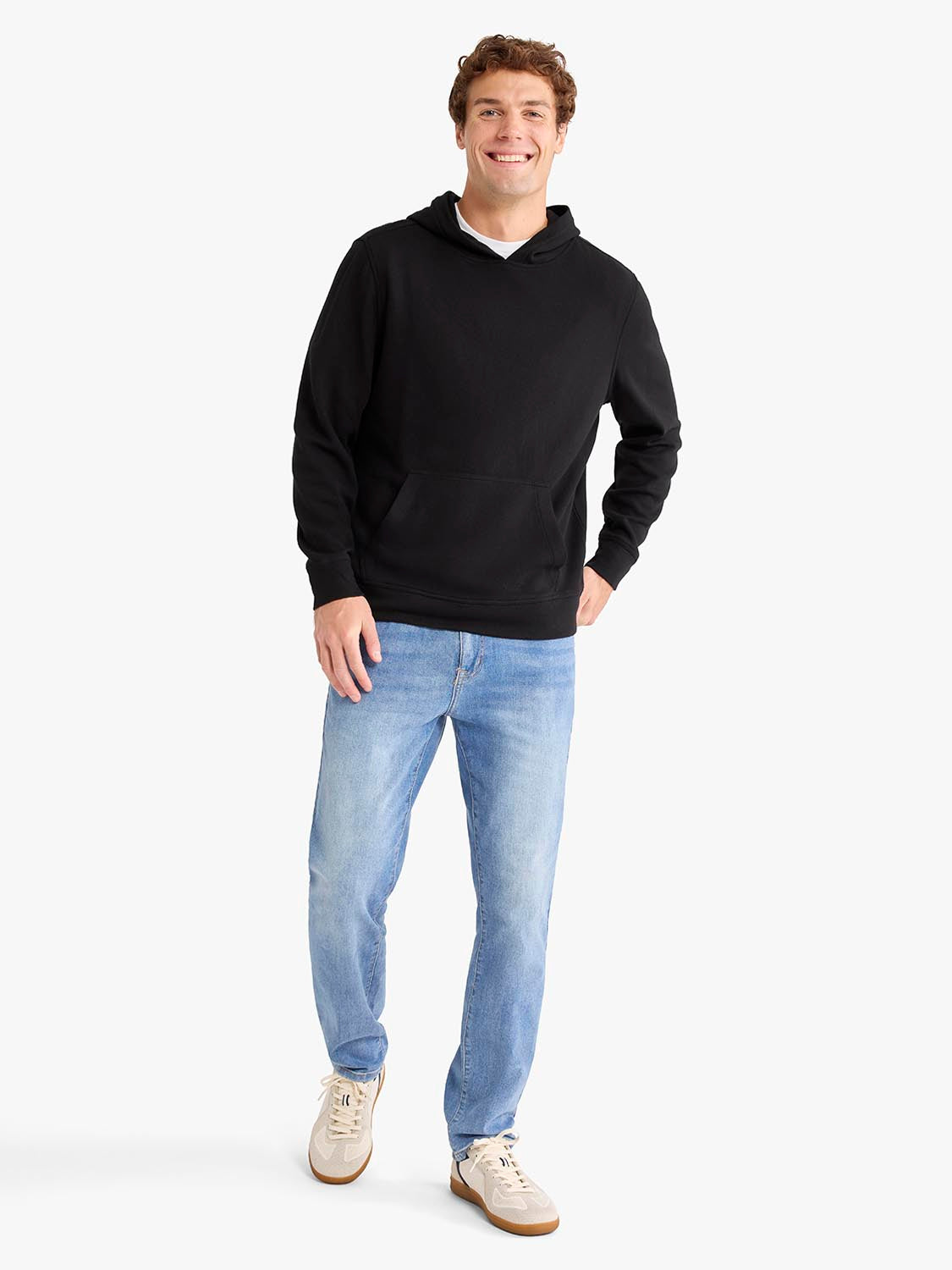 Man wearing a black hoodie with front pocket and blue jeans standing against white background.