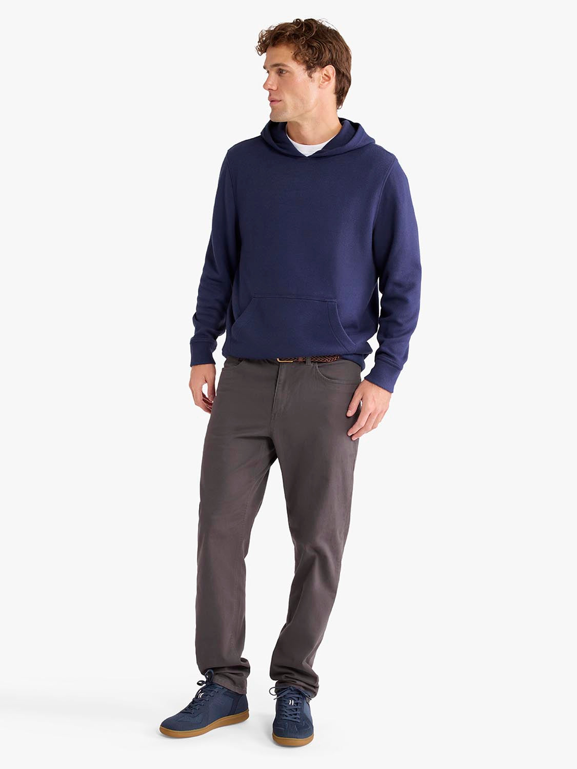 Man wearing a navy hoodie with a kangaroo pocket and gray pants with navy shoes.
