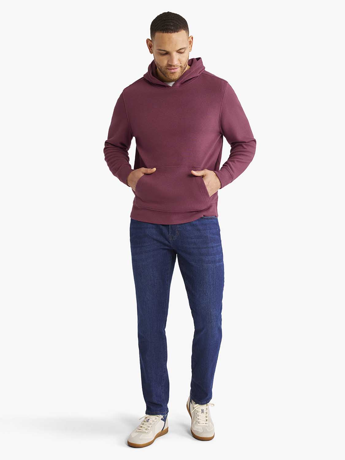 Man wearing a burgundy hoodie with front kangaroo pockets and dark jeans with cream sneakers.