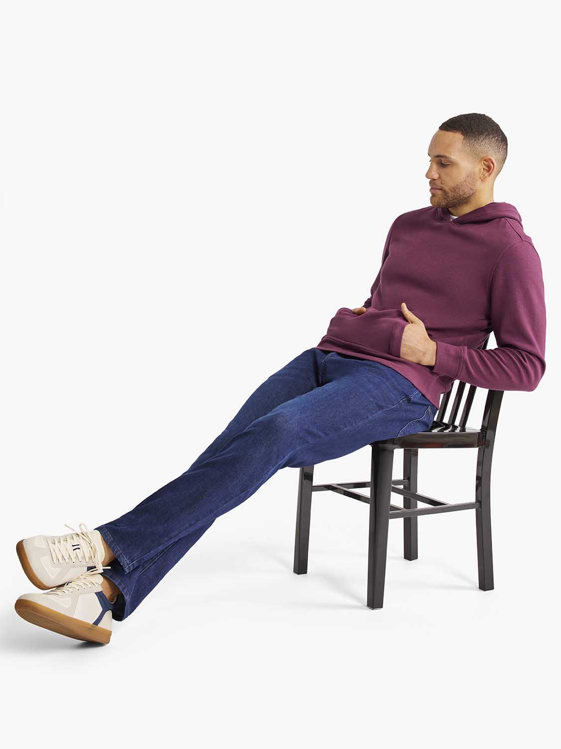 Man seated on a wooden chair wearing a burgundy hoodie and dark blue jeans with cream sneakers.