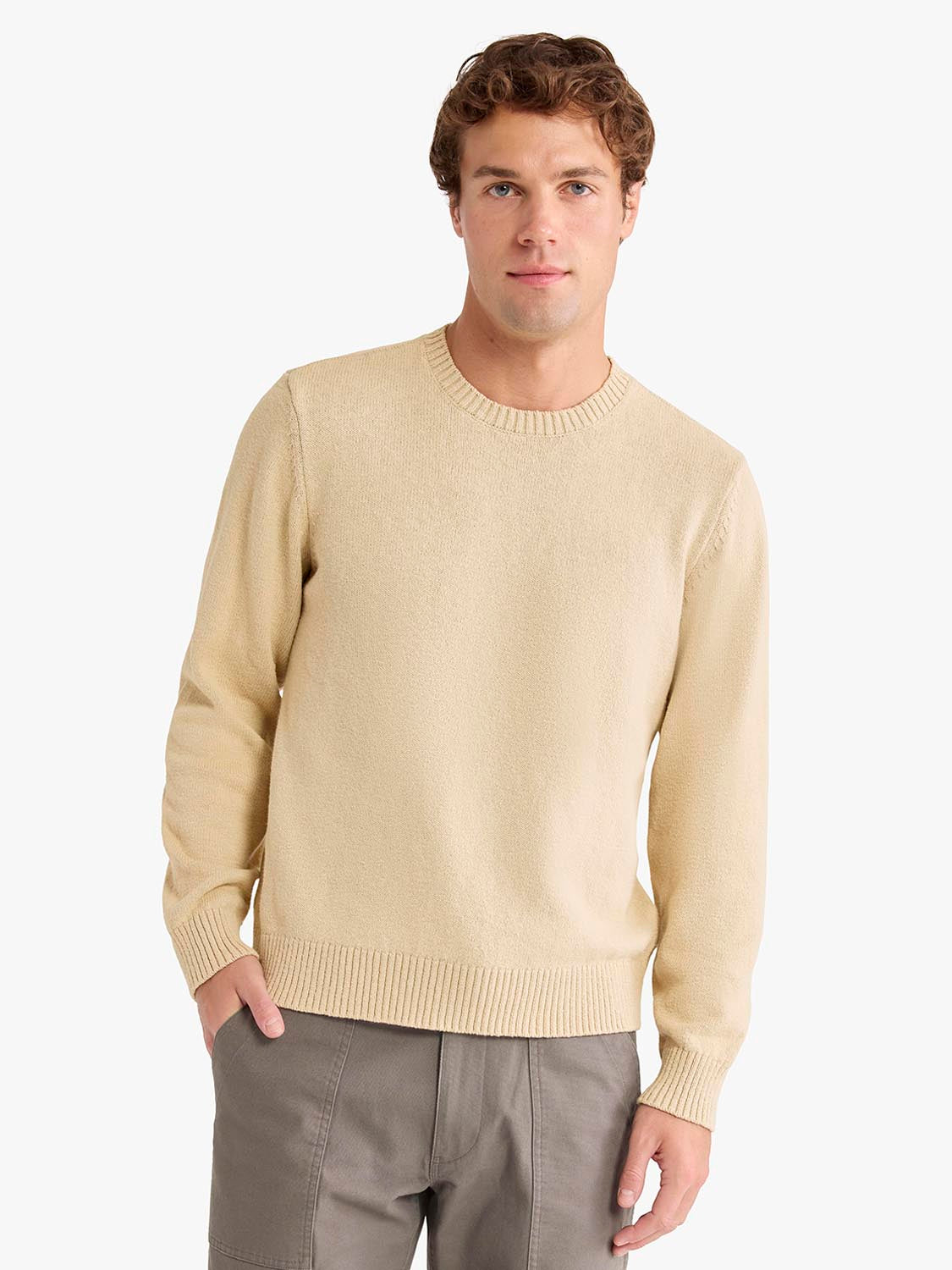 Man wearing a beige crew neck sweater with ribbed cuffs and hem over gray pants.