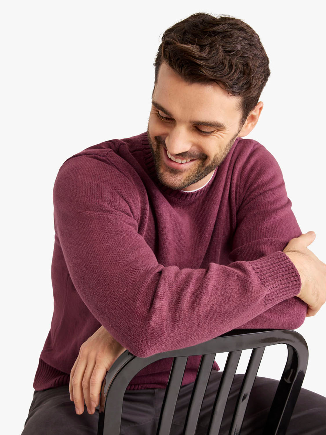 Man wearing a burgundy crew neck sweater with ribbed sleeves and cuffs, seated on a chair.
