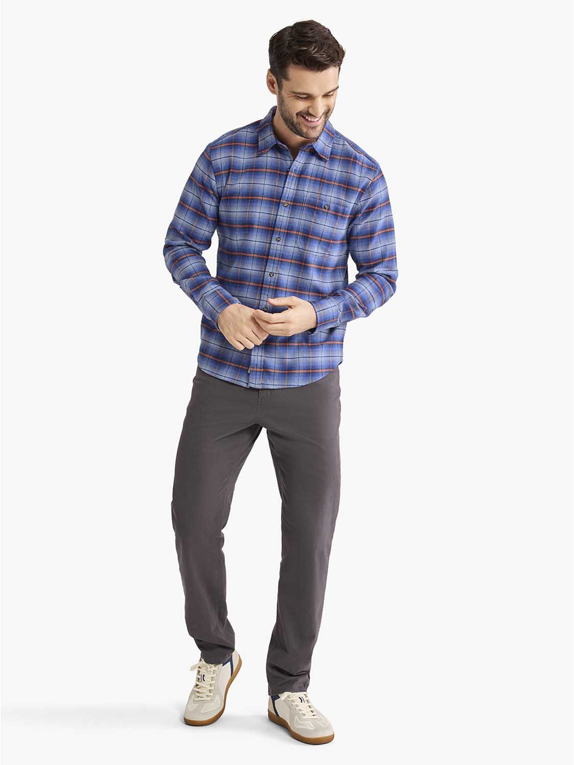 Man wearing a blue plaid flannel shirt with long sleeves and gray pants standing against a white background.